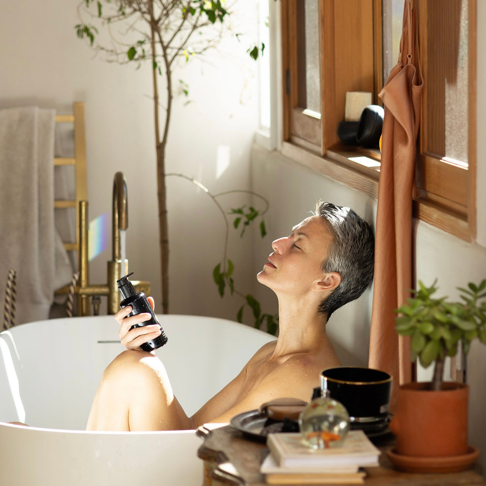 Woman in a bathtub holding a bottle, surrounded by plants and a cup.